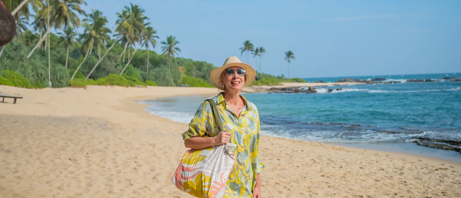 A smiling woman in a yellow dress and hat walks along a sandy beach with palm trees and ocean in the background.