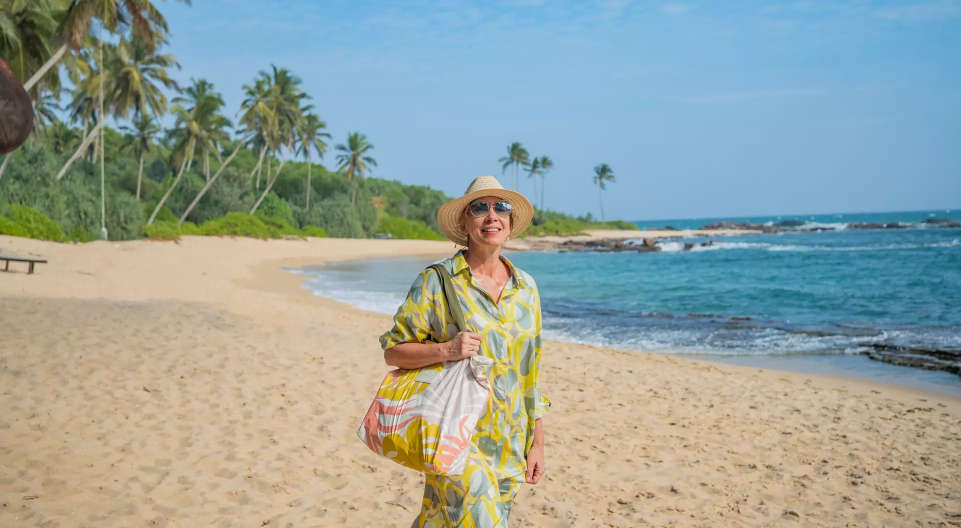 A smiling woman in a yellow dress and hat walks along a sandy beach with palm trees and ocean in the background.