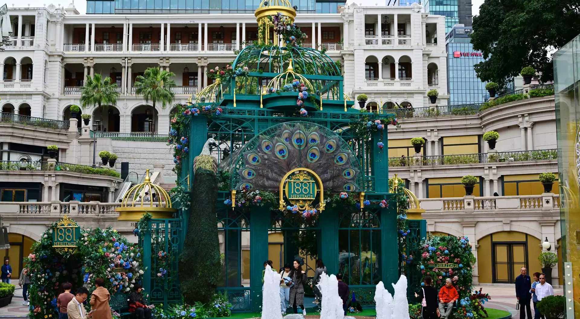A colorful structure with peacock design and fountains, surrounded by greenery and buildings in a bustling plaza.