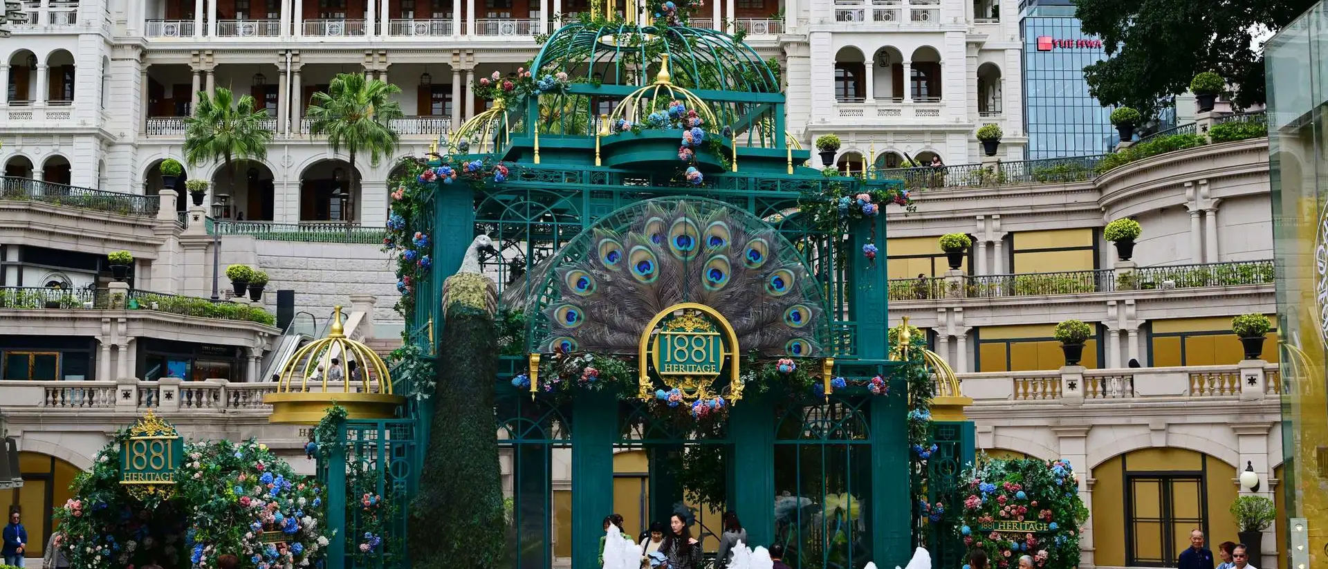 A colorful structure with peacock design and fountains, surrounded by greenery and buildings in a bustling plaza.