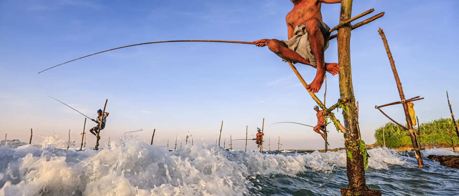A fisherman balances on a wooden pole over waves, holding a fishing rod against a scenic coastal backdrop.