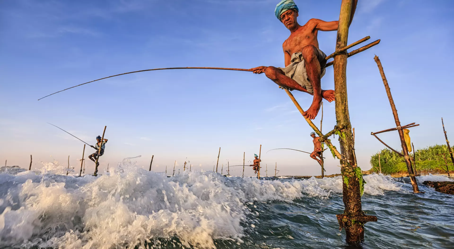 A fisherman balances on a wooden pole over waves, holding a fishing rod against a scenic coastal backdrop.