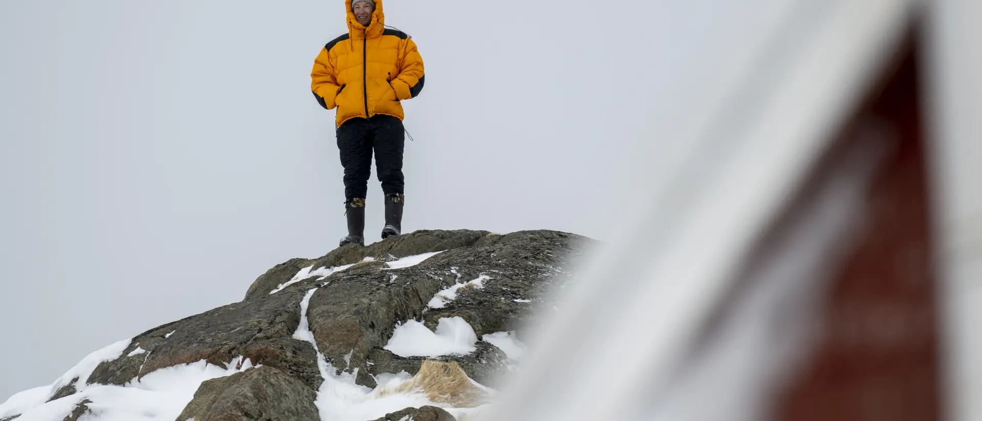 A person in a bright yellow jacket stands on a snowy rock, overlooking a winter landscape with a blurred structure nearby.