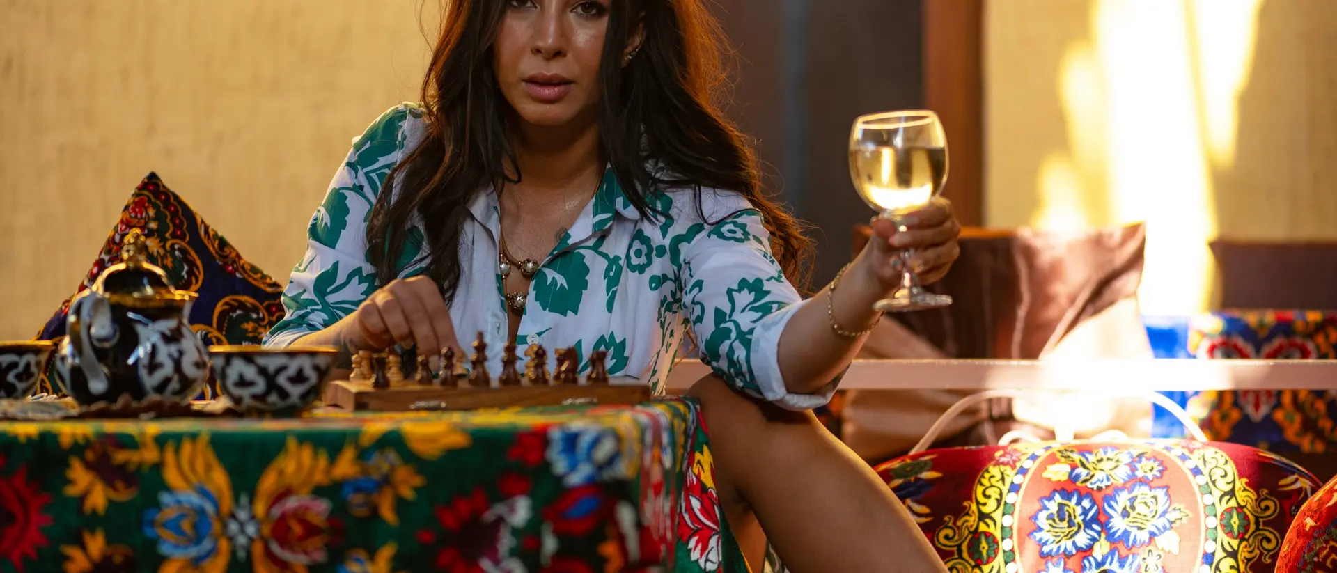 A woman with long hair sits at a colorful table, holding a glass of wine while playing chess in a vibrant setting.