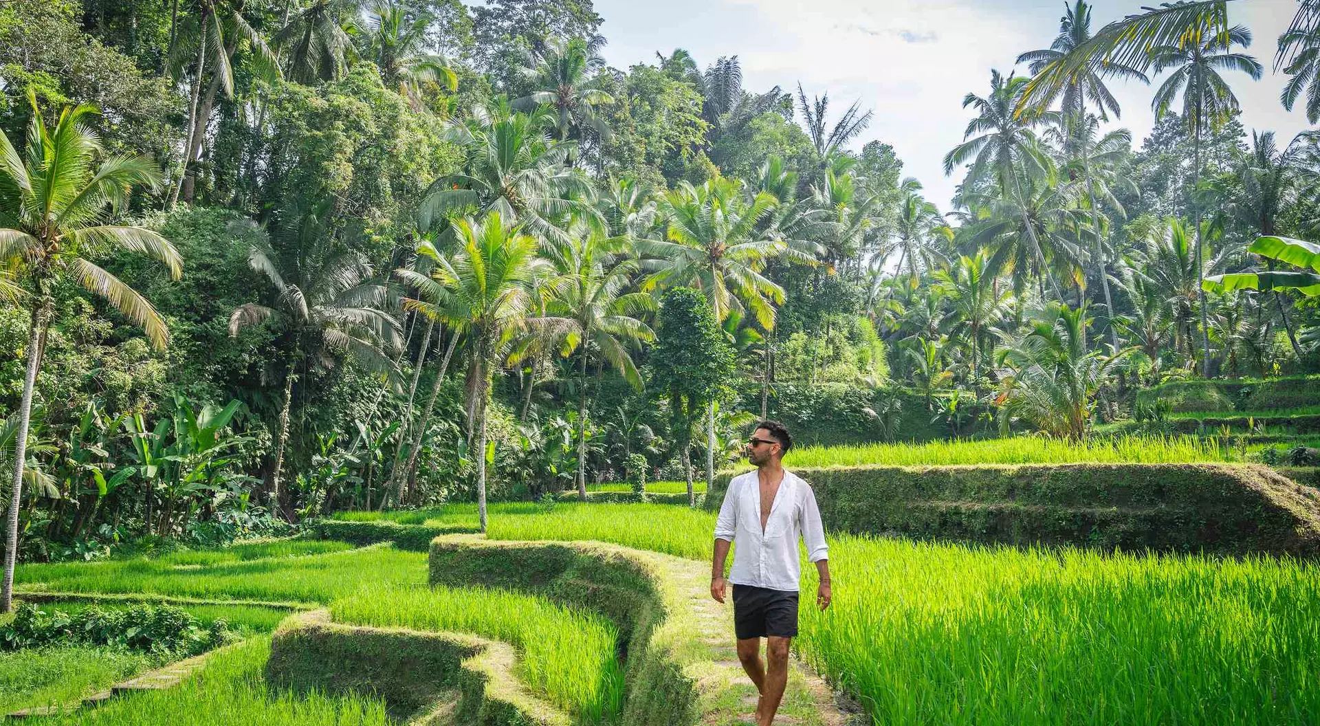 A person walking barefoot along a path through lush green rice terraces surrounded by tropical trees.