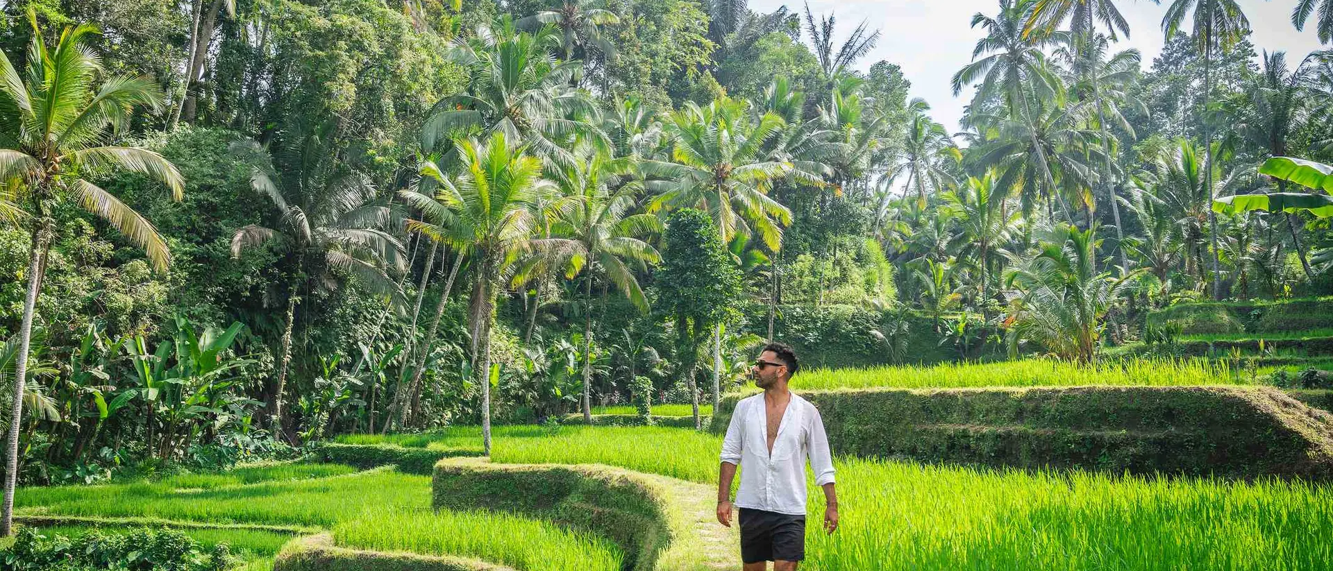 A person walking barefoot along a path through lush green rice terraces surrounded by tropical trees.