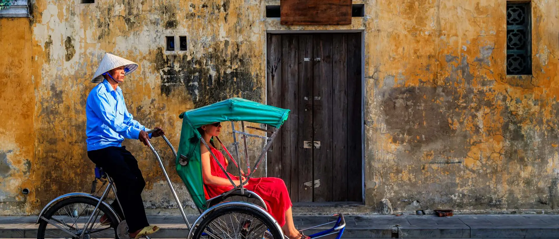 A rickshaw driver in a conical hat transports a woman in a red dress against a weathered yellow wall.