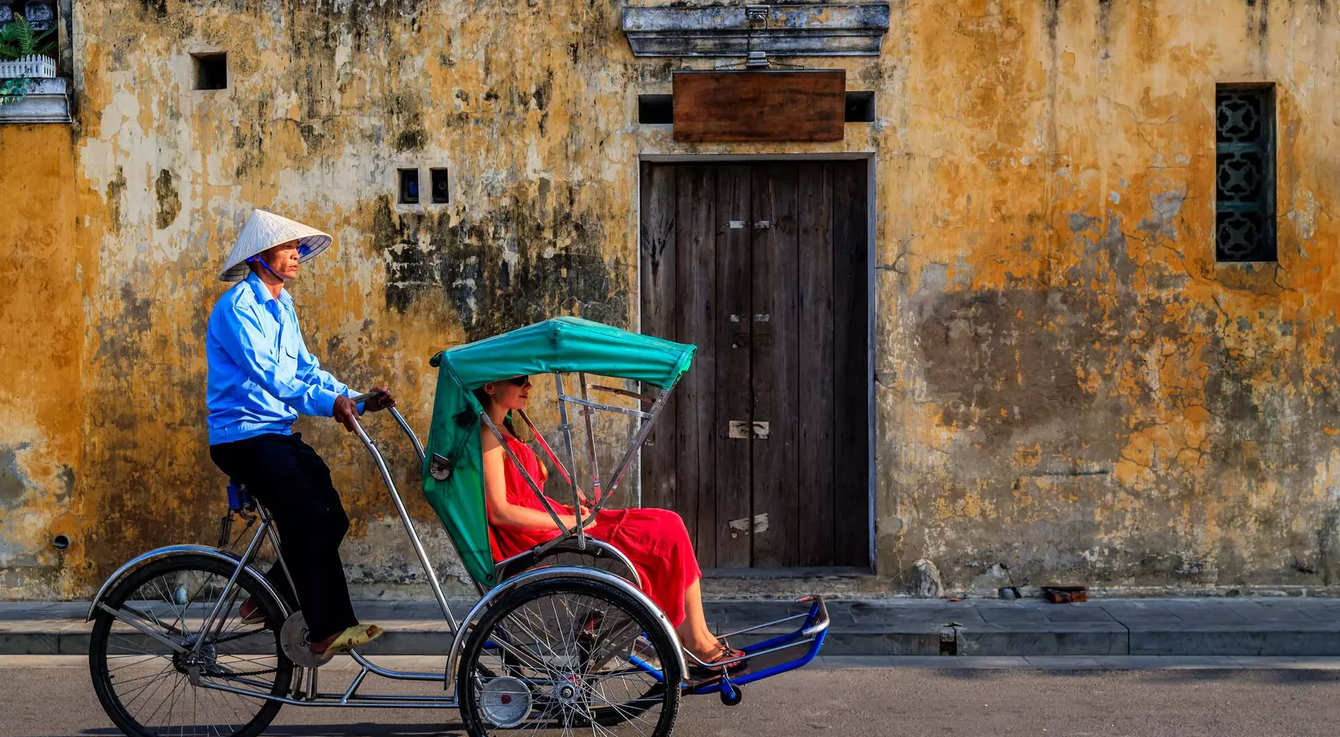 A rickshaw driver in a conical hat transports a woman in a red dress against a weathered yellow wall.