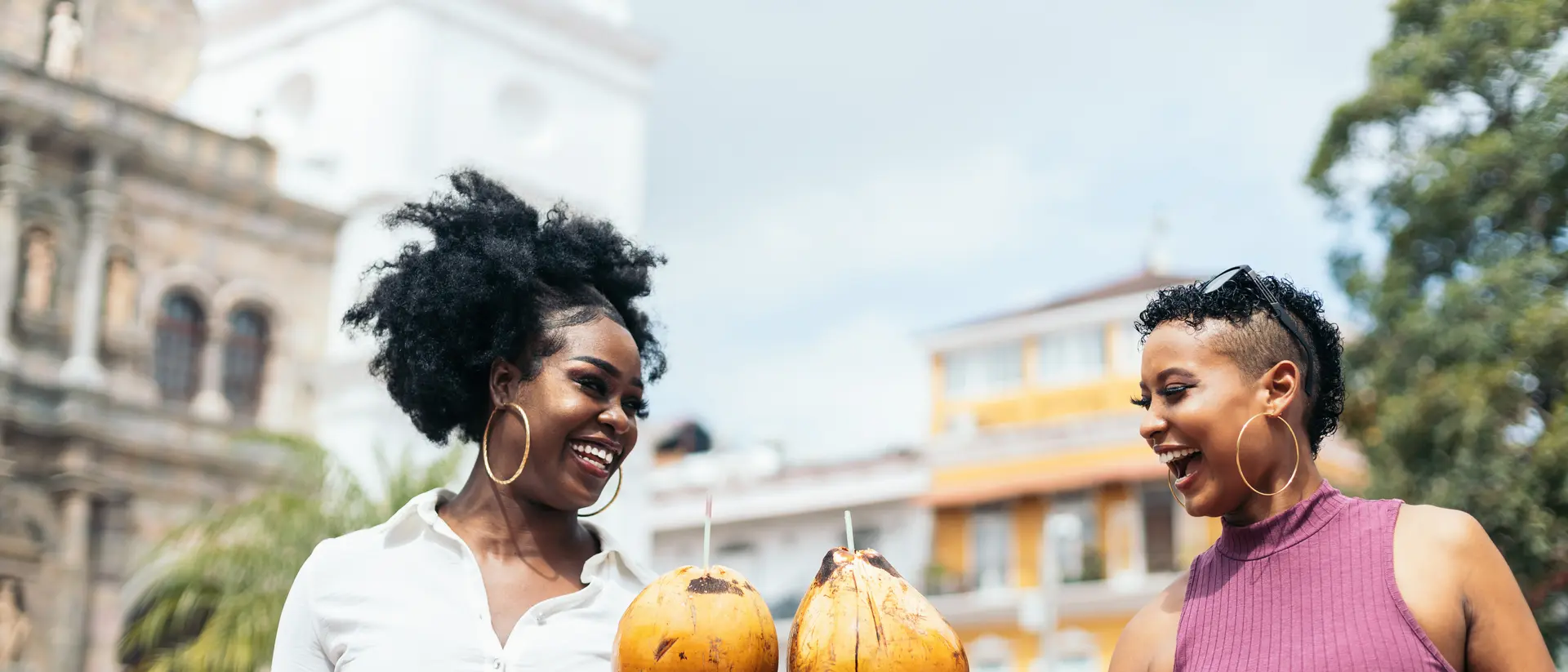 Two women smile and hold coconuts in a vibrant outdoor setting with historic buildings and greenery in the background.