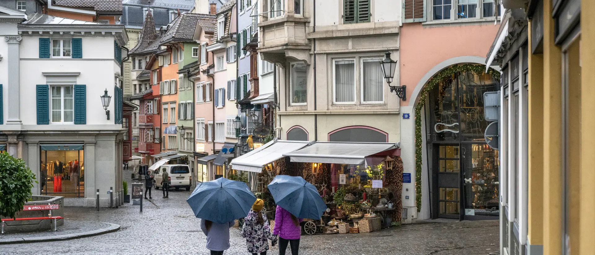 Three women with umbrellas walk down a cobblestone street lined with colorful buildings and shops on a rainy day.