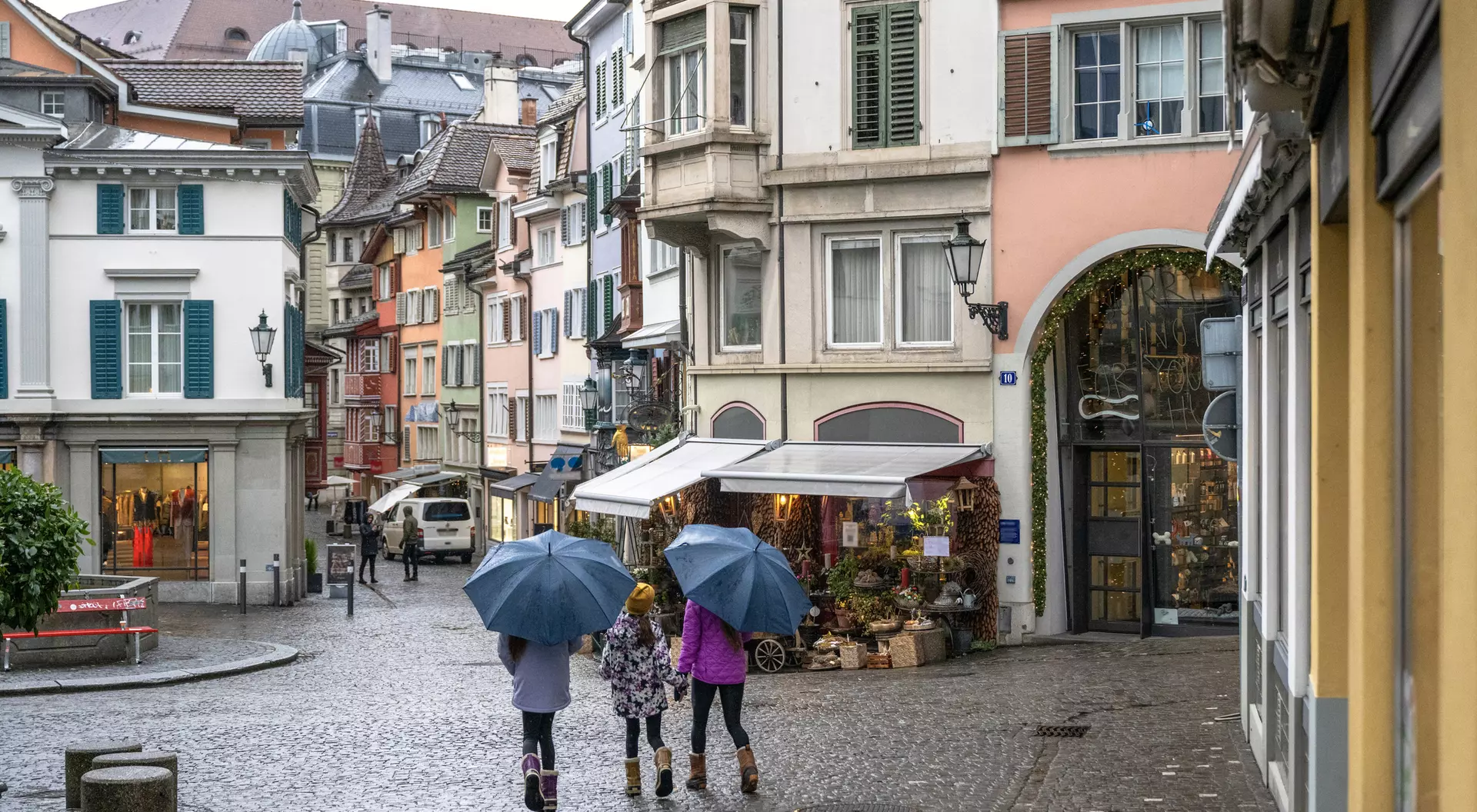 Three women with umbrellas walk down a cobblestone street lined with colorful buildings and shops on a rainy day.