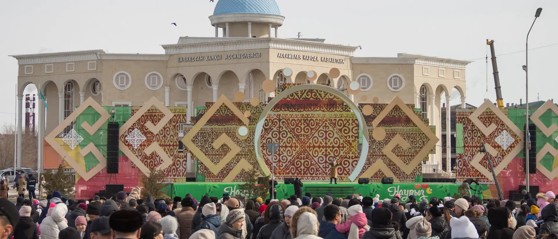 Crowd gathered in front of a large building with decorative patterns for a public event outdoors on a clear day.