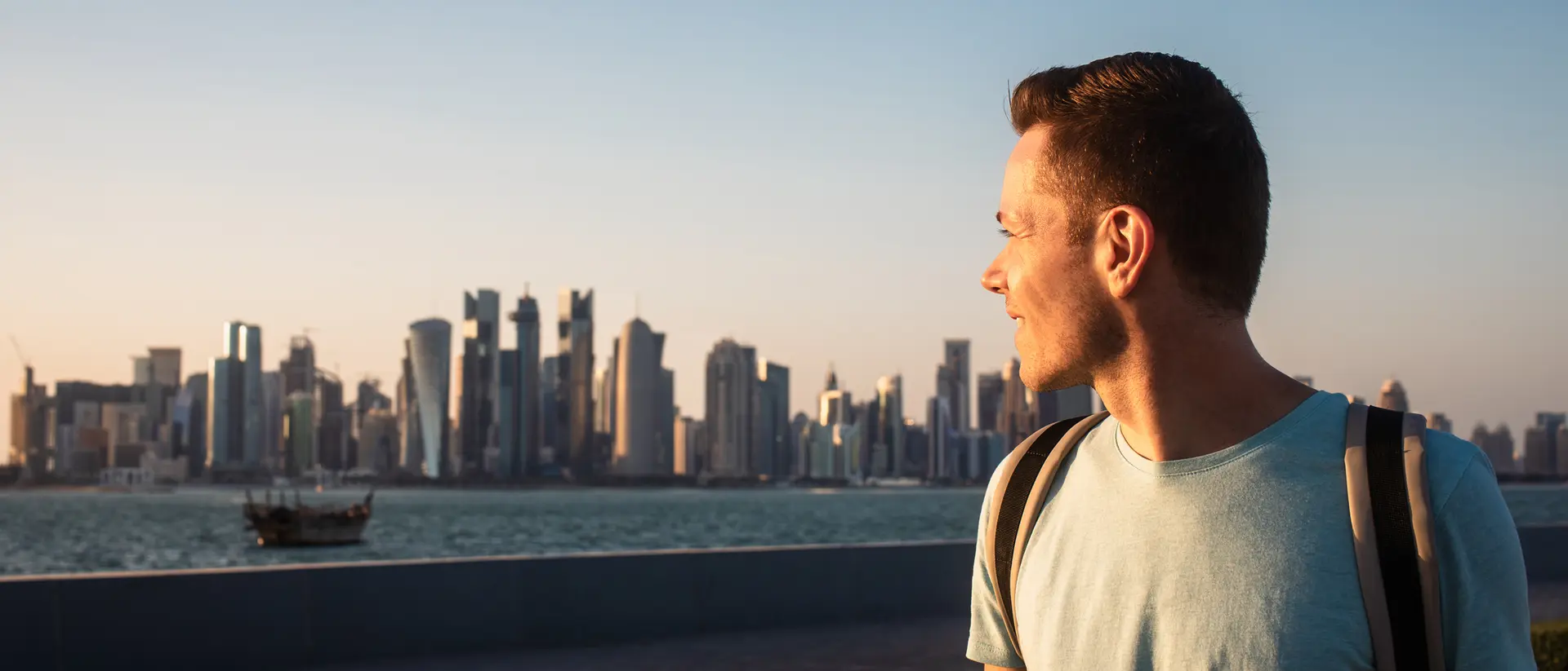 A man gazes at a city skyline during sunset, with water and a boat in the foreground.