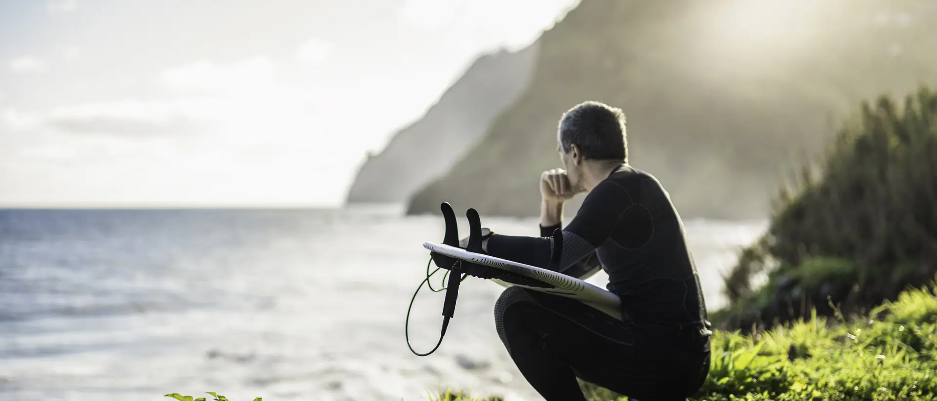 A person in a wetsuit sits on a grassy cliff, gazing at the ocean and mountains under a bright sky.