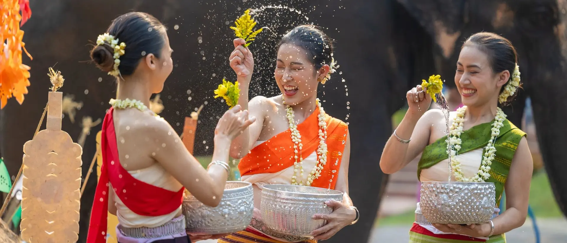Three women in colorful traditional outfits joyfully splash water while celebrating with an elephant in the background.