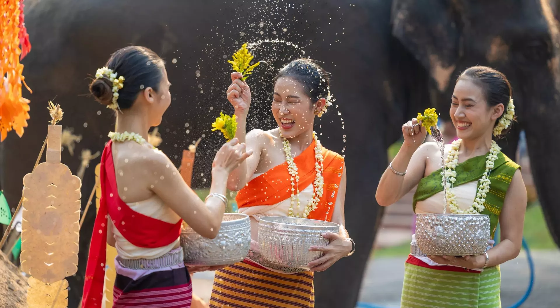 Three women in colorful traditional outfits joyfully splash water while celebrating with an elephant in the background.