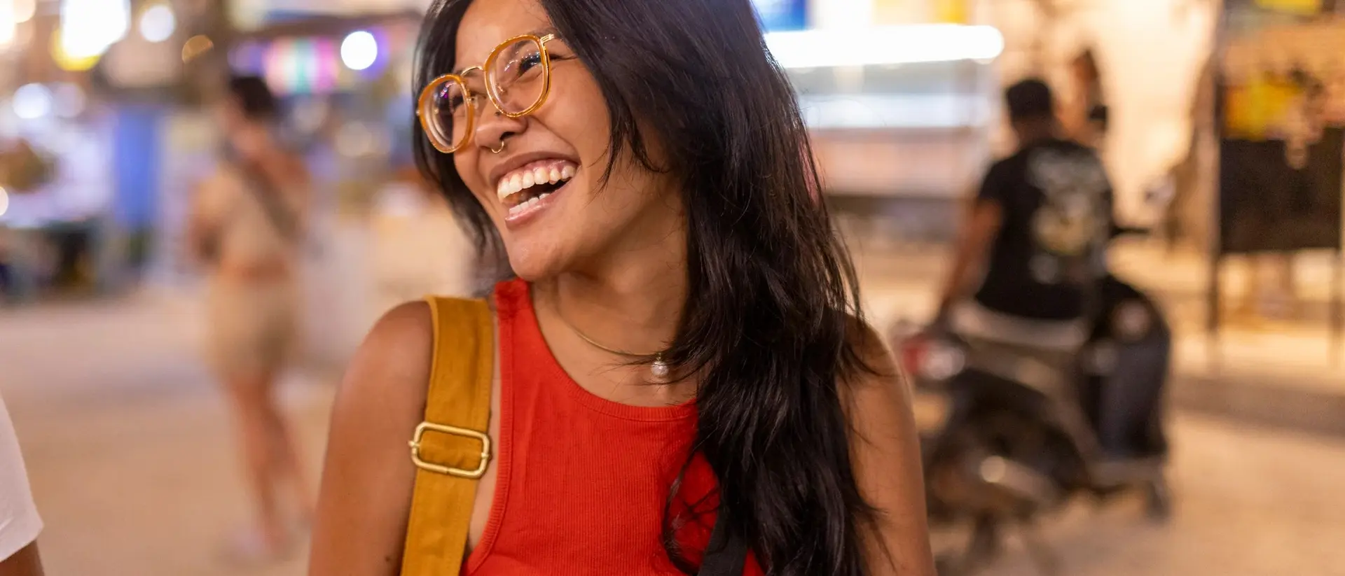Smiling woman with long black hair, wearing glasses and a red shirt, standing in a bustling outdoor setting.