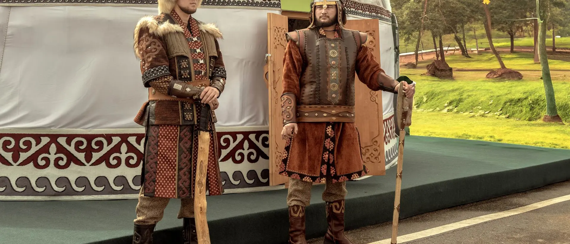 Two men in traditional clothing stand outside a yurt, holding staffs, with a green landscape in the background.