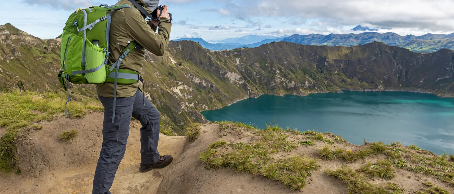 Hiker in green backpack photographing a scenic mountain lake surrounded by hills under a partly cloudy sky.