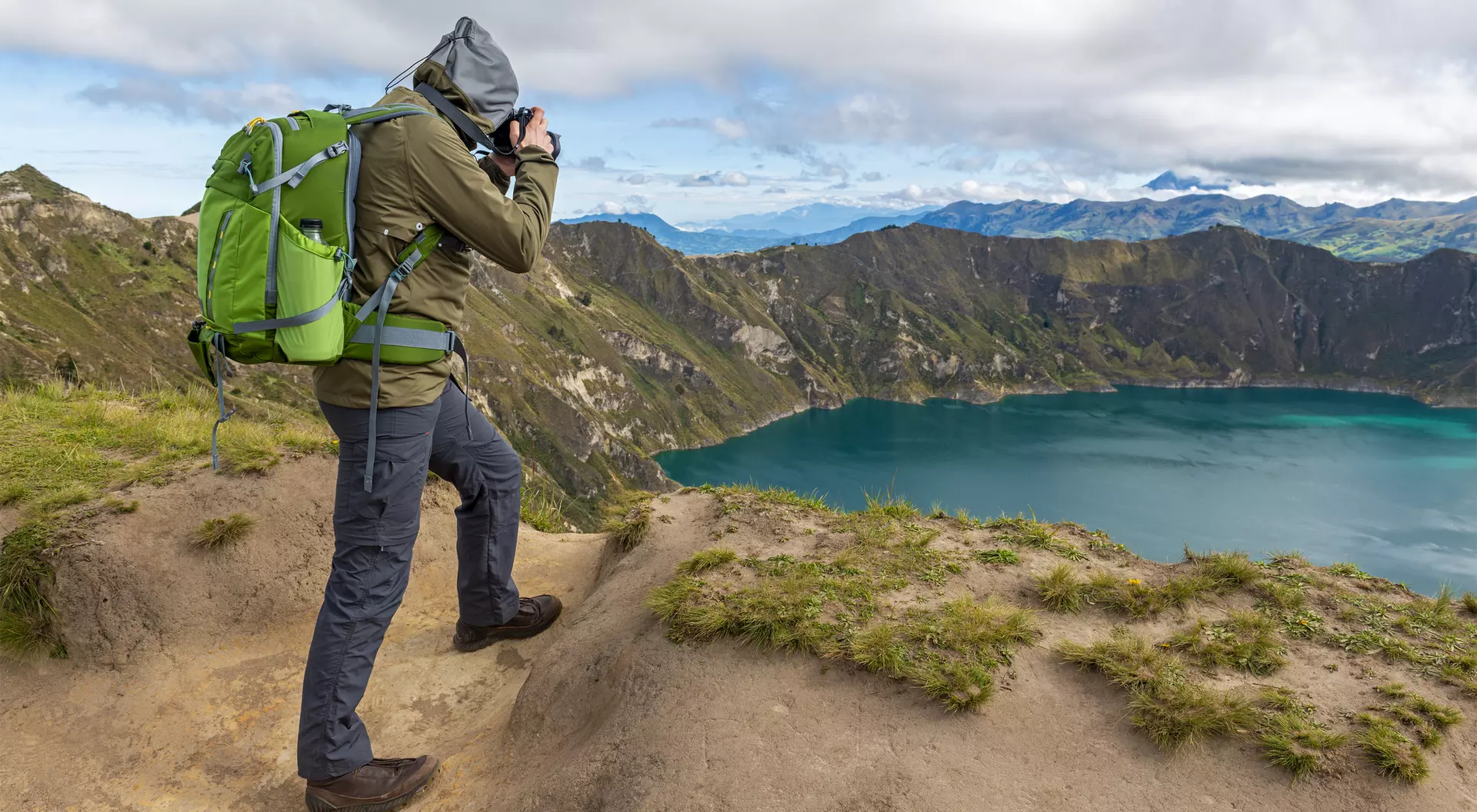 Hiker in green backpack photographing a scenic mountain lake surrounded by hills under a partly cloudy sky.