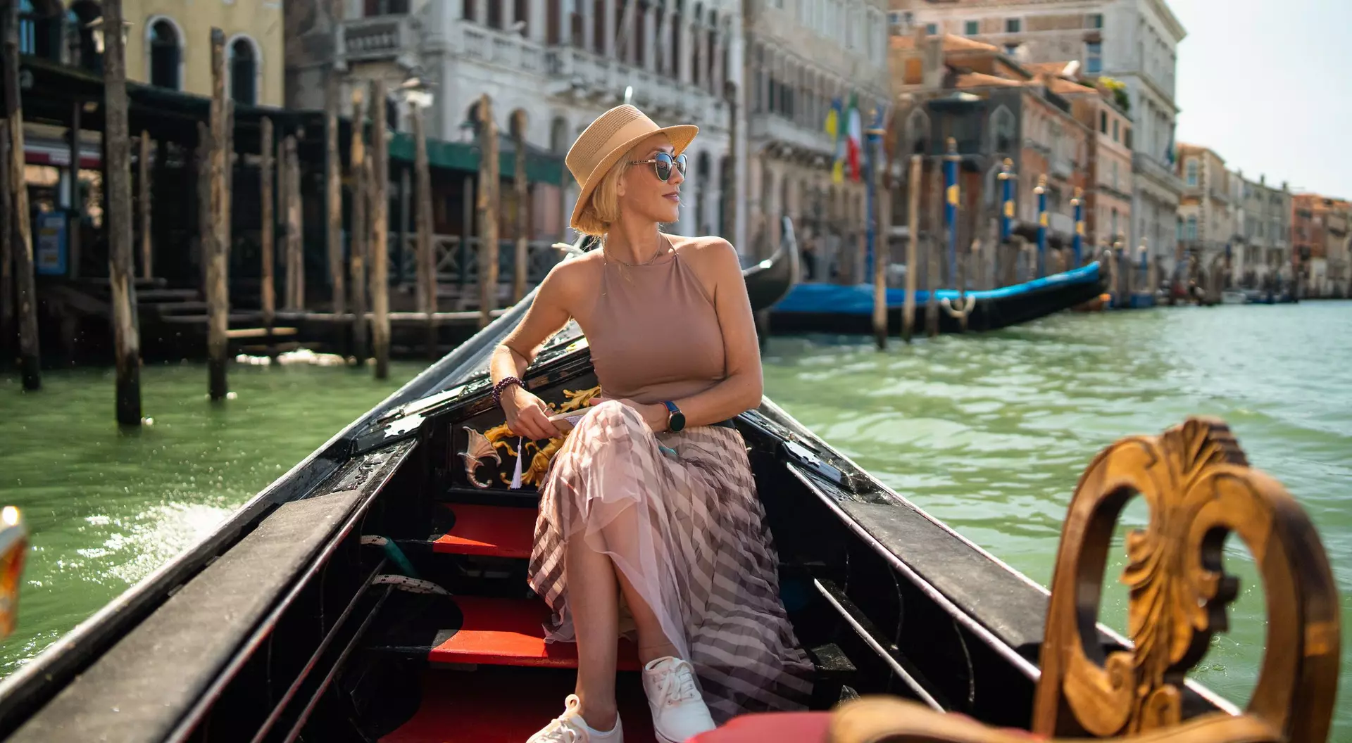 Woman wearing a hat and sunglasses relaxes in a gondola on a canal, with buildings and boats in the background.