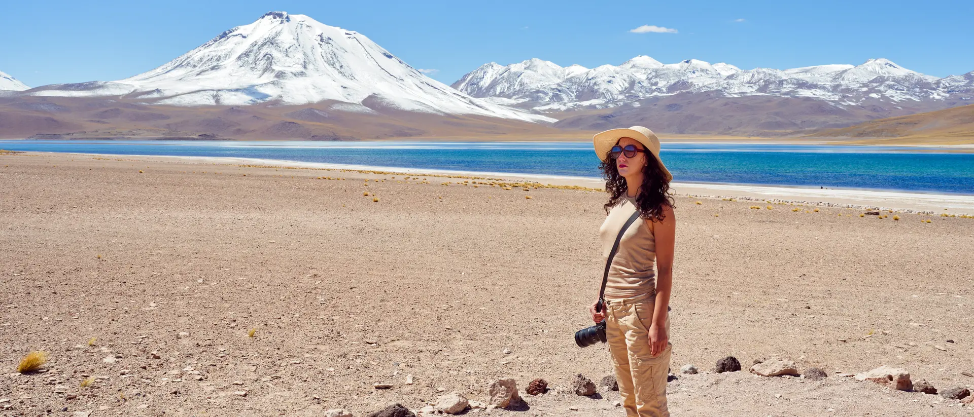 A person stands on a rocky shore by a turquoise lake, with a snow-capped mountain in the background under a clear blue sky.
