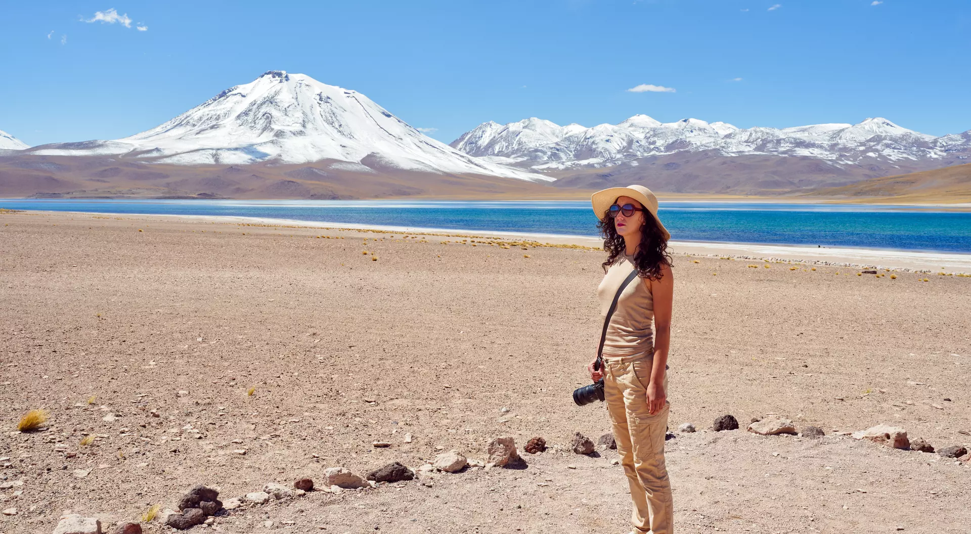 A person stands on a rocky shore by a turquoise lake, with a snow-capped mountain in the background under a clear blue sky.