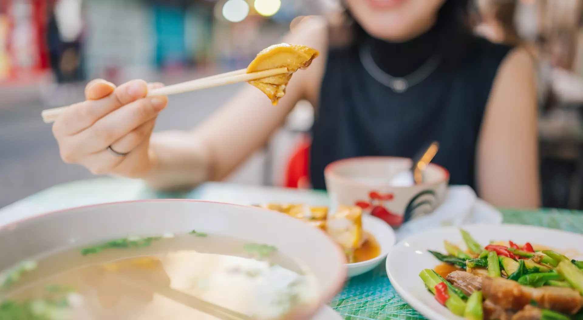 A person holds a piece of food with chopsticks, smiling at a table with a bowl of soup and colorful dishes.
