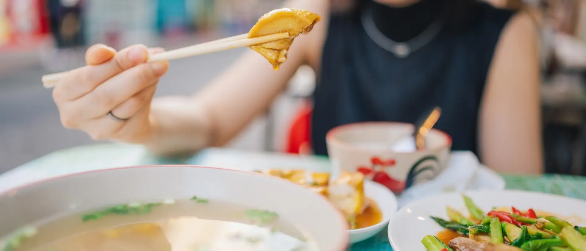 A person holds a piece of food with chopsticks, smiling at a table with a bowl of soup and colorful dishes.