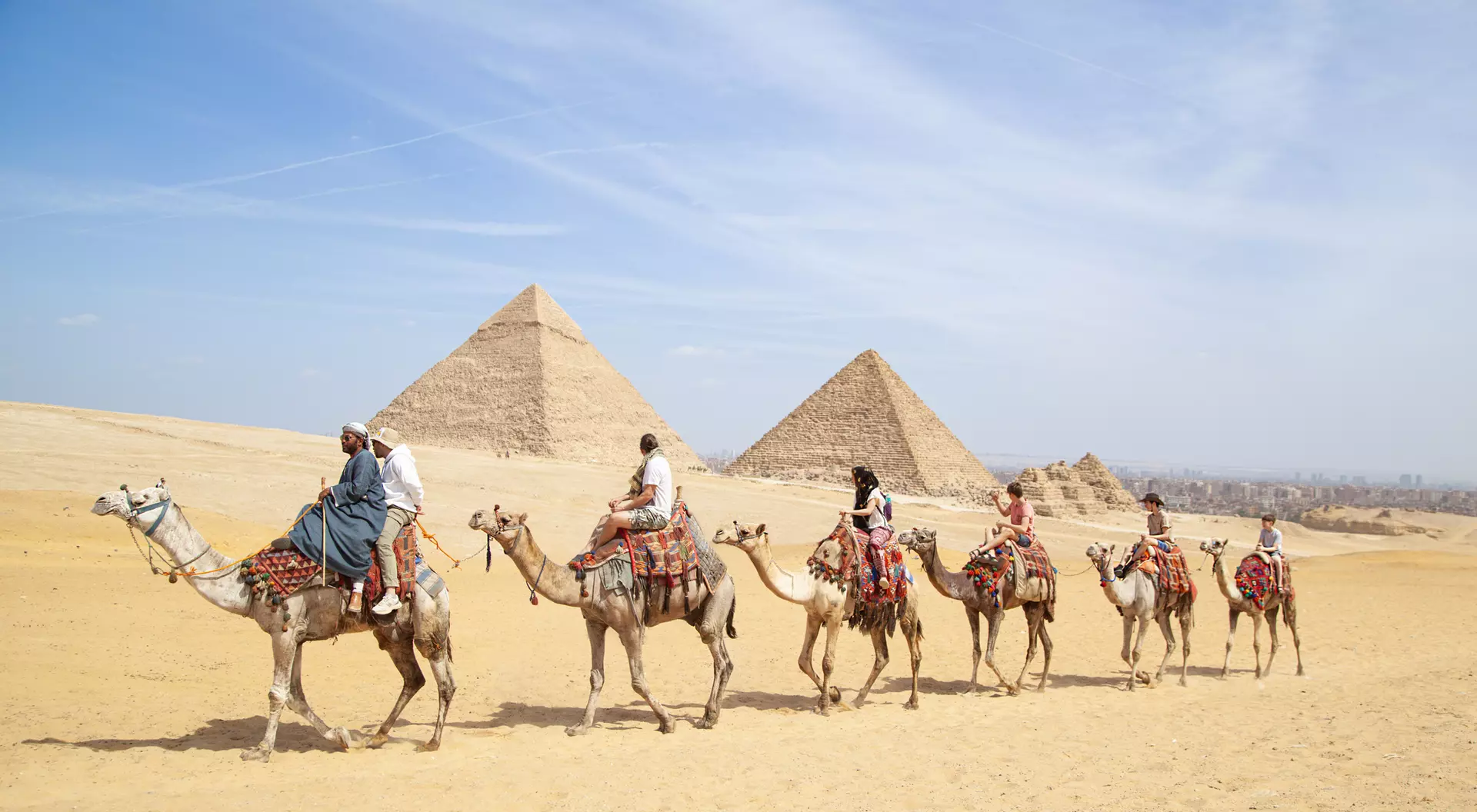 A group of people riding camels in the desert near the pyramids of Giza under a clear blue sky.