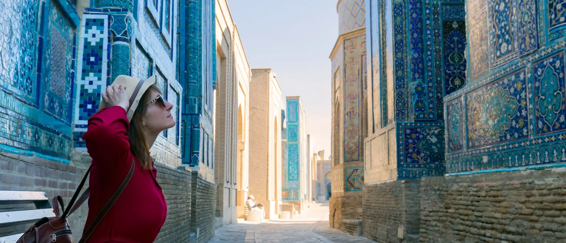 A woman in a red dress and hat sits on a bench in a narrow, blue-tiled alleyway, looking up at the architecture.