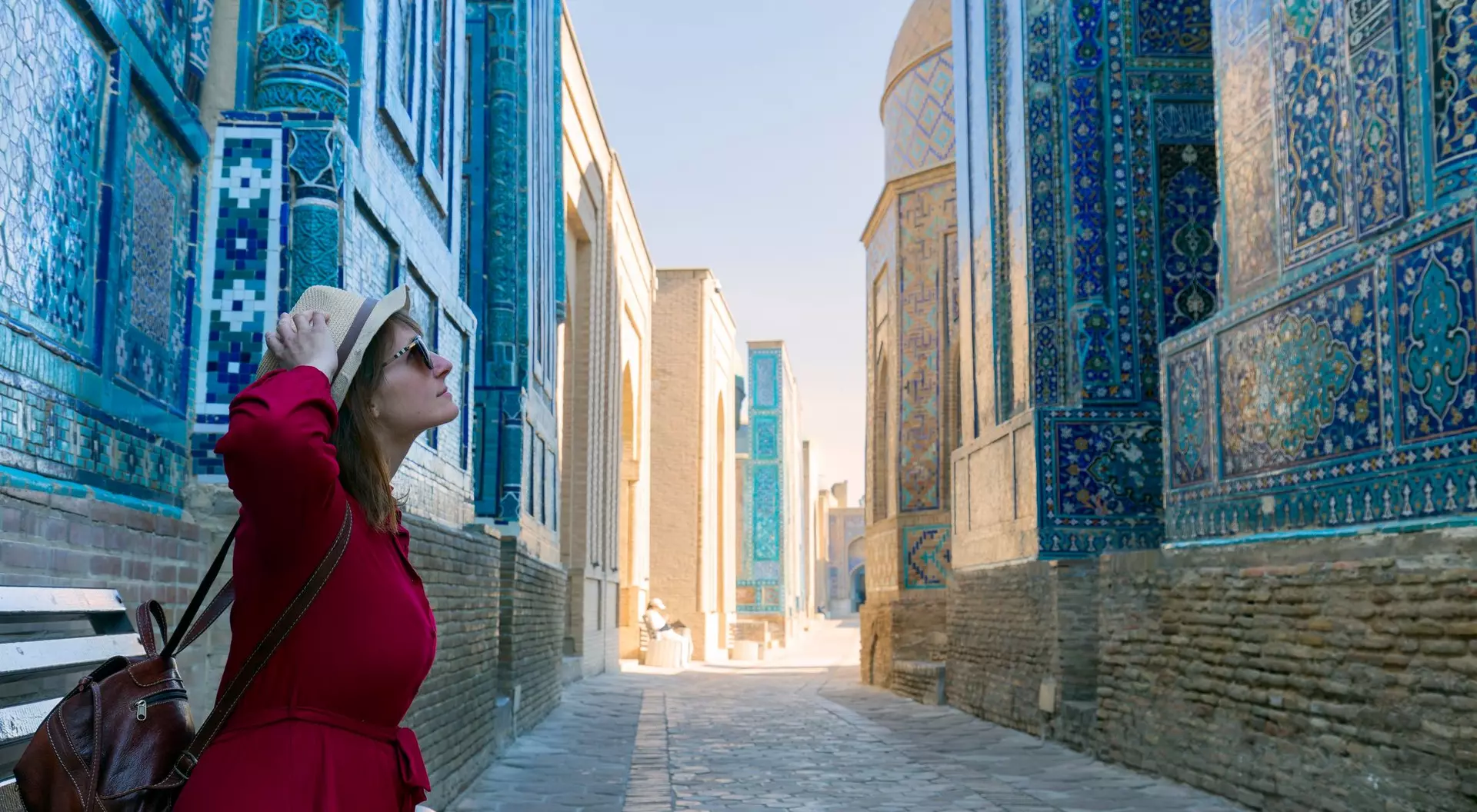 A woman in a red dress and hat sits on a bench in a narrow, blue-tiled alleyway, looking up at the architecture.