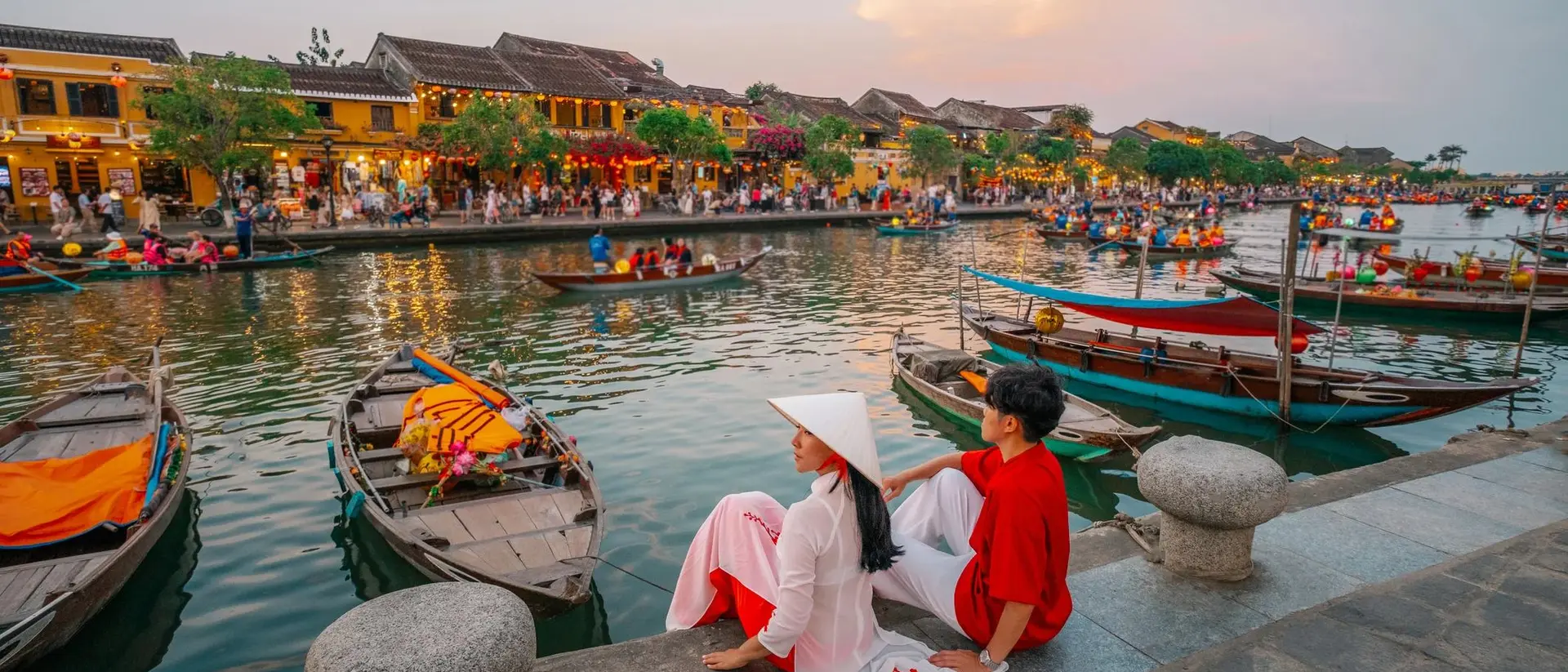 A couple sits by a riverbank in Hoi An, Vietnam, surrounded by boats and lively market scenes at sunset.