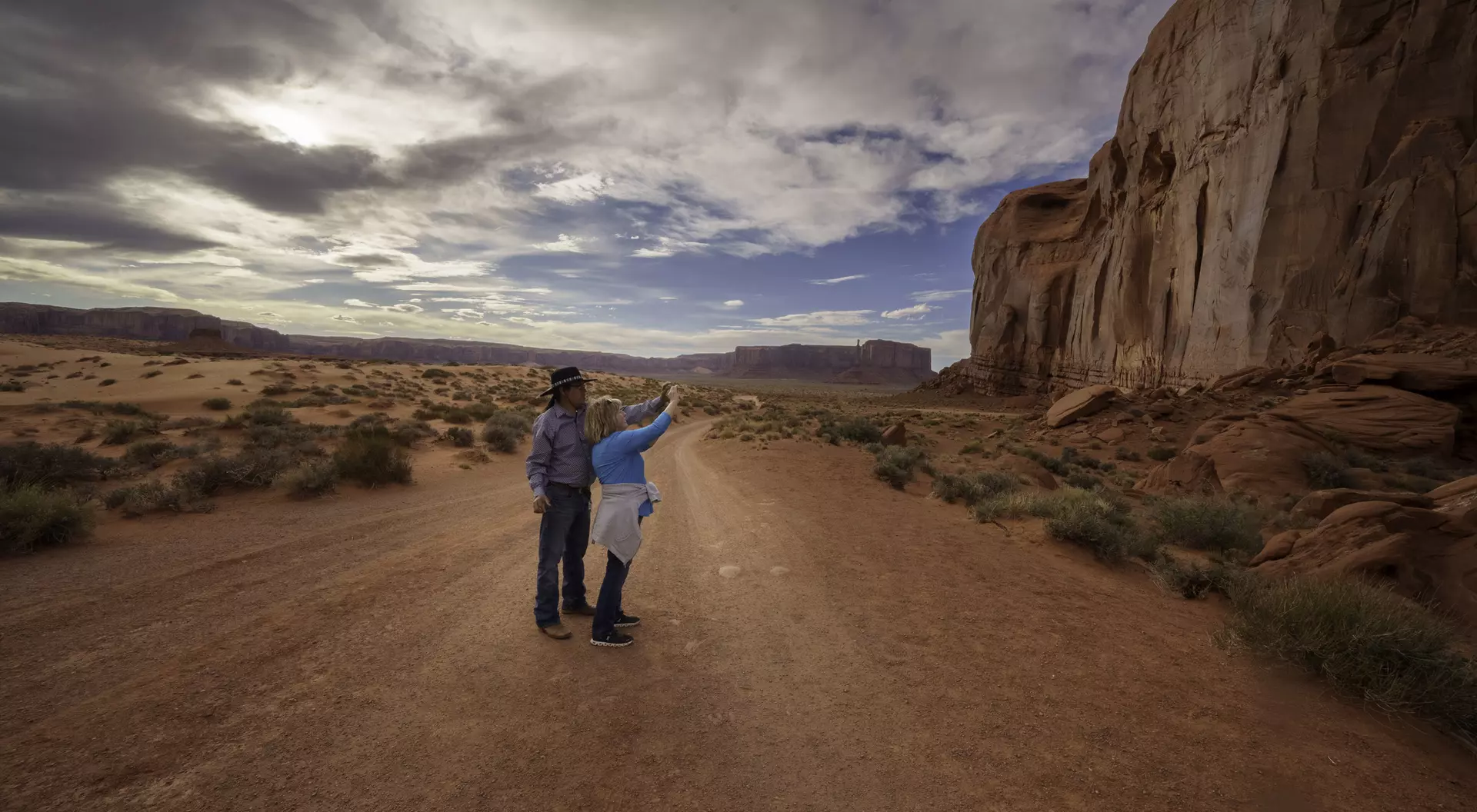 Two people stand on a dirt road, pointing at a large rock formation under a cloudy sky in a desert landscape.