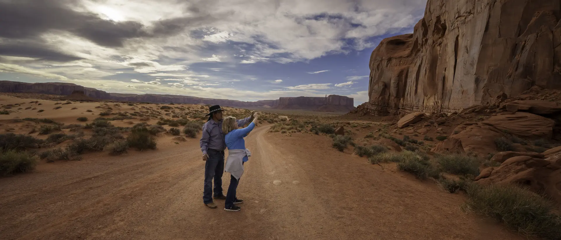 Two people stand on a dirt road, pointing at a large rock formation under a cloudy sky in a desert landscape.