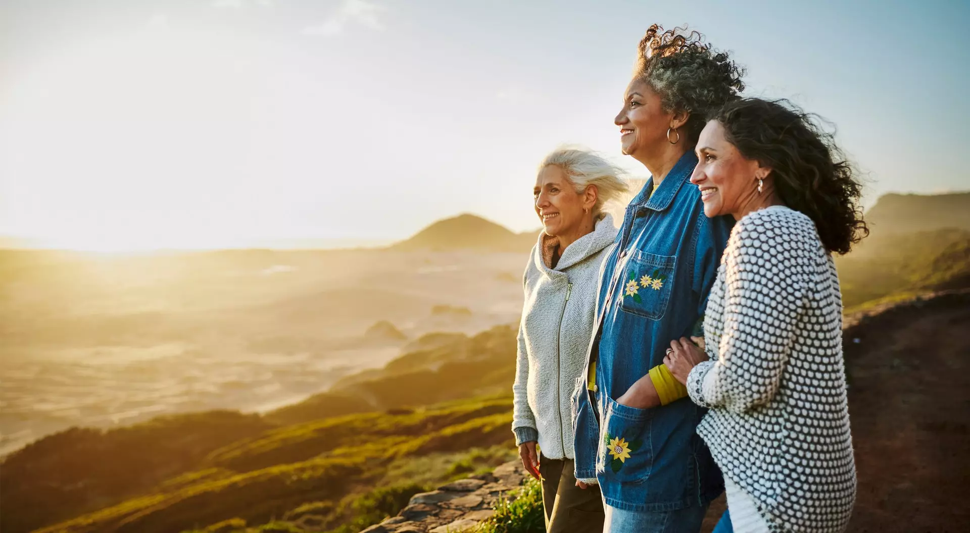 Three smiling older women stand together on a hillside, facing the sun with a landscape of hills and ocean in the background.