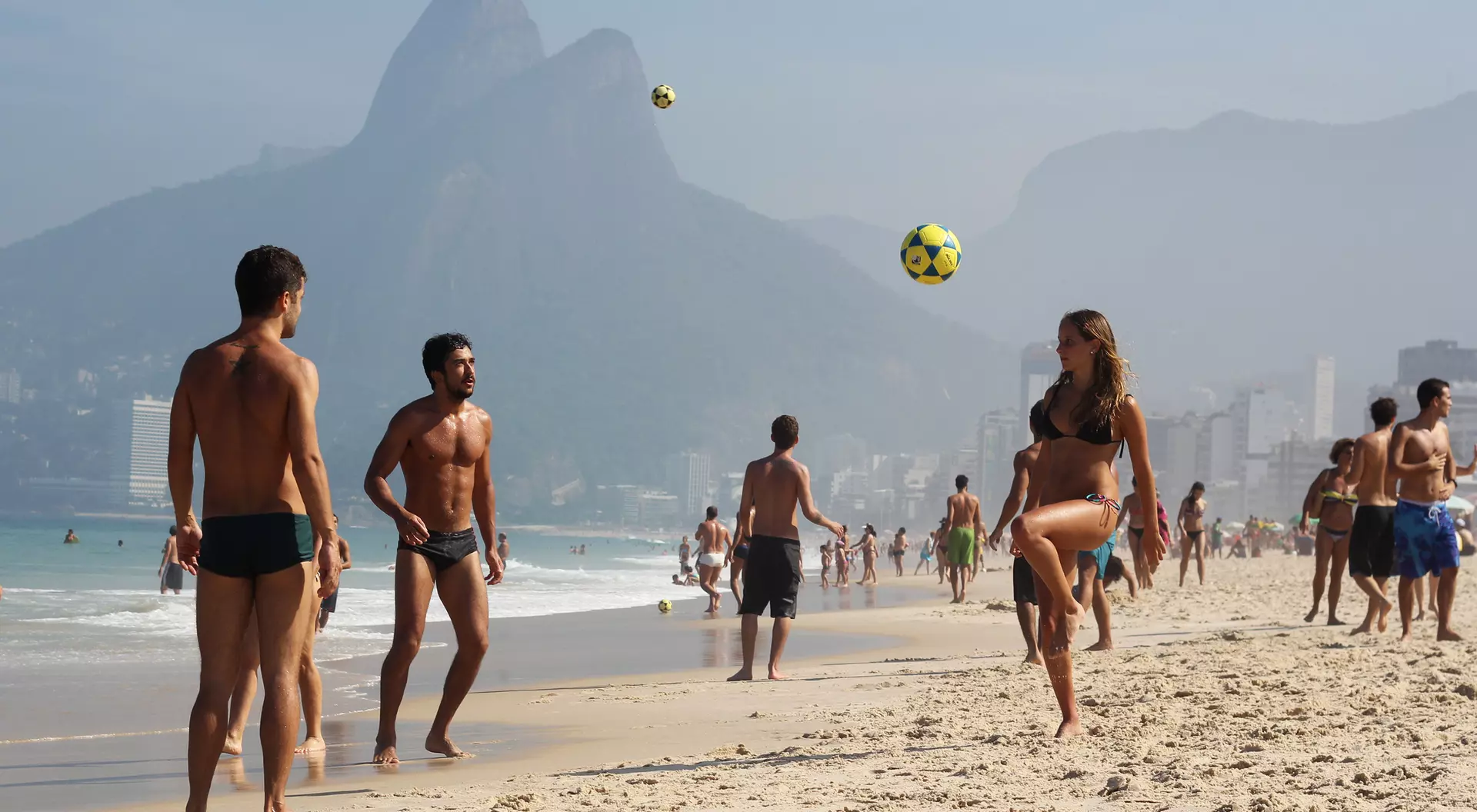 A sandy beach scene with people playing soccer, mountains in the background, and a clear blue sky.