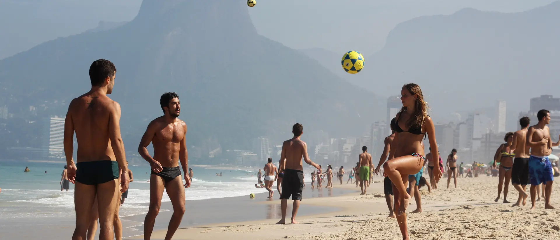 A sandy beach scene with people playing soccer, mountains in the background, and a clear blue sky.