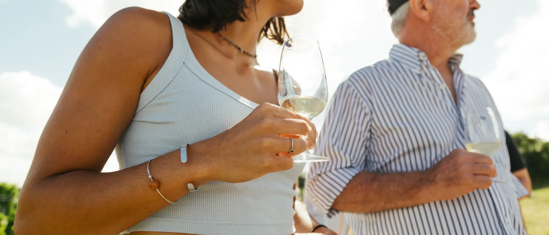 A woman holding a wine glass stands next to an older man, both enjoying a sunny vineyard setting.