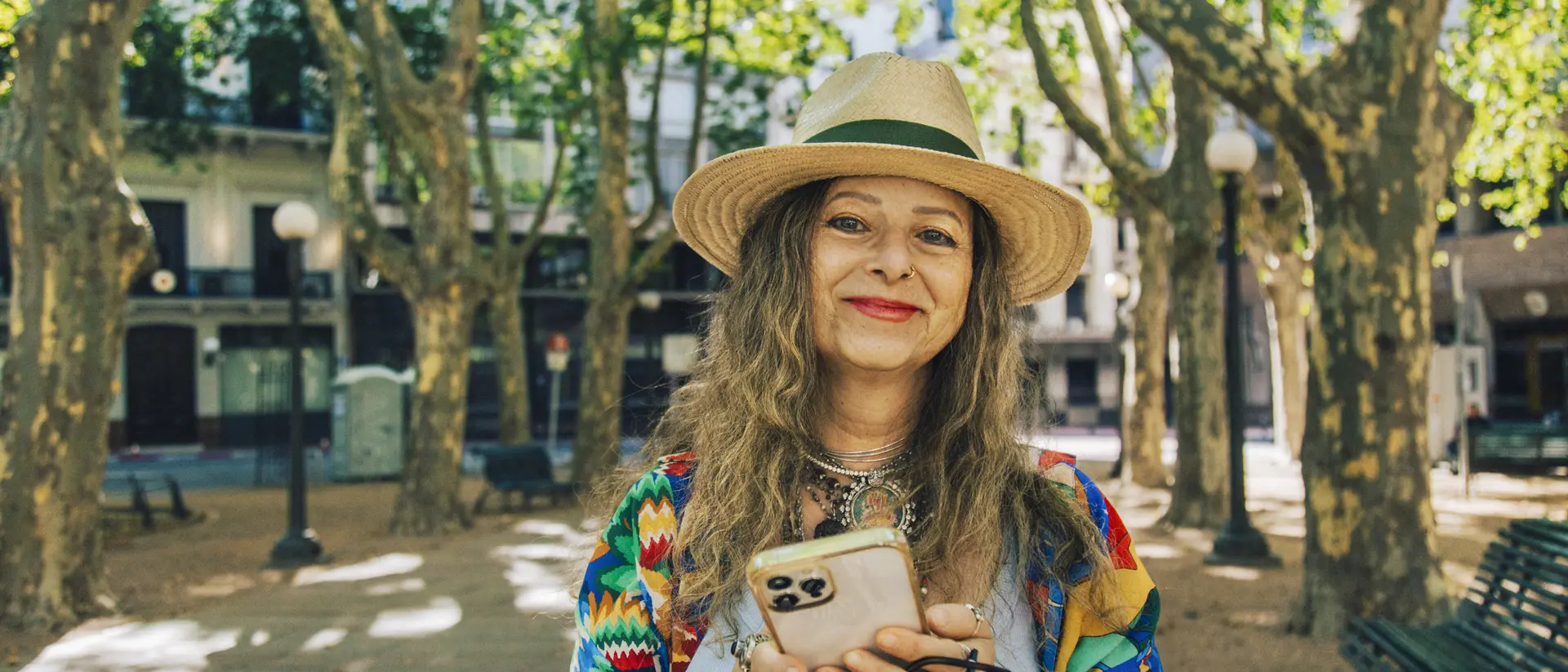 Smiling woman with long, wavy hair, wearing a straw hat and colorful clothing, holding a phone in a sunny park setting.