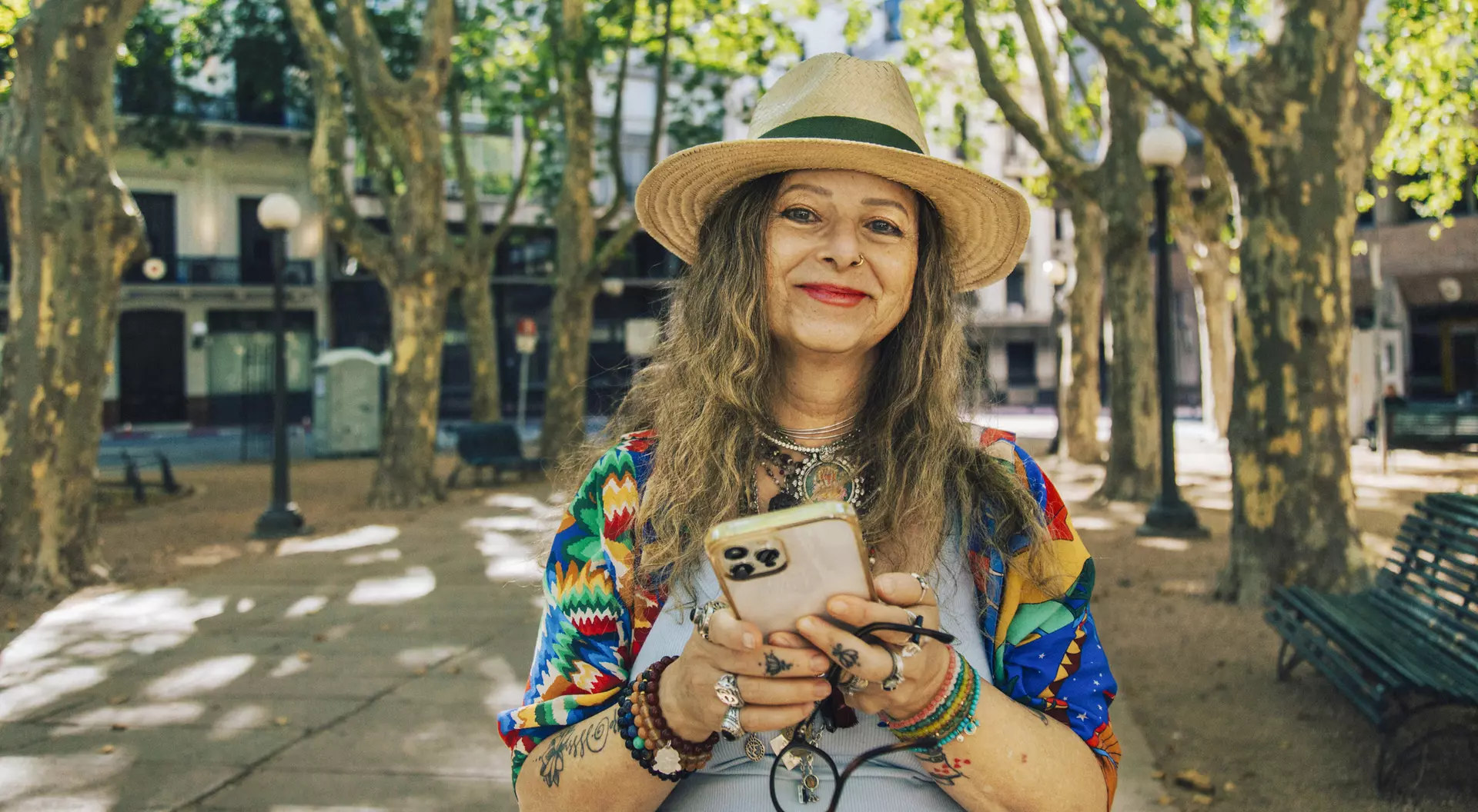 Smiling woman with long, wavy hair, wearing a straw hat and colorful clothing, holding a phone in a sunny park setting.