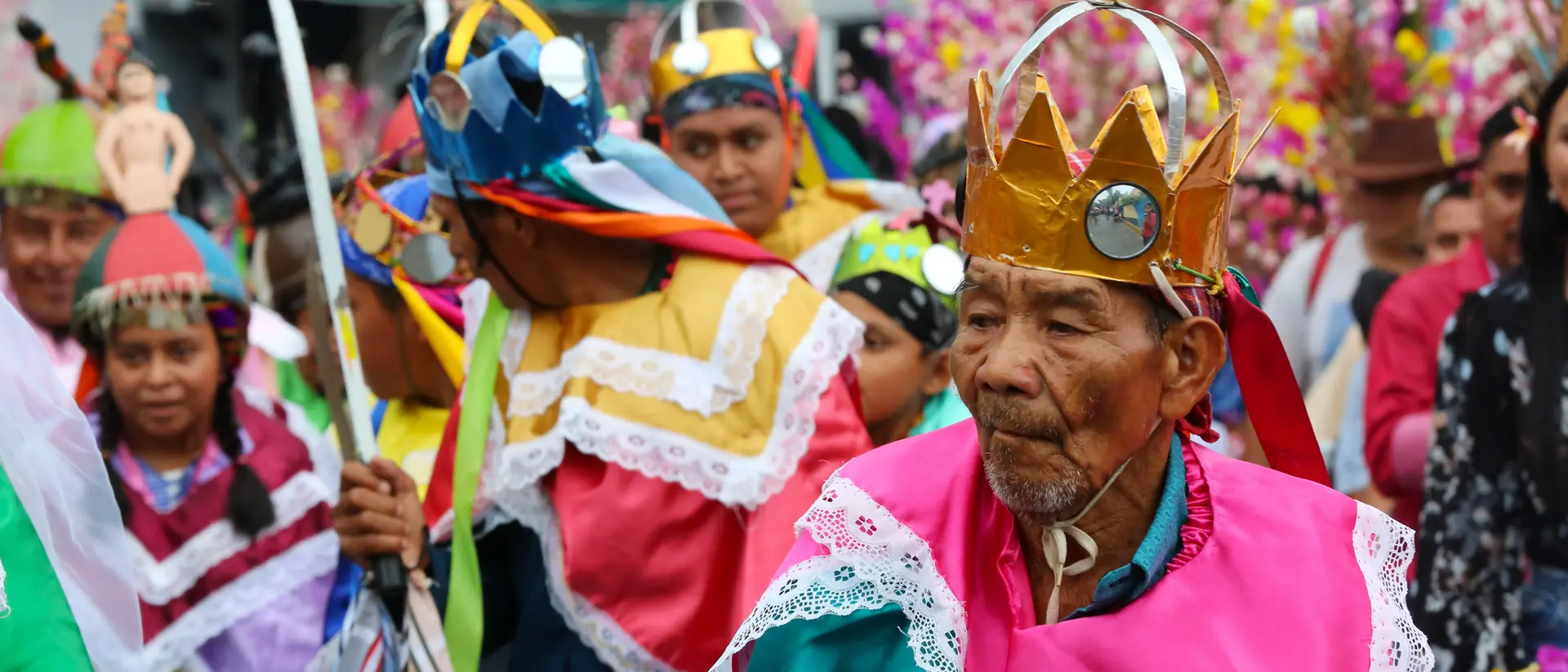 A vibrant parade featuring people in colorful costumes and crowns, celebrating with joyful expressions amidst festive decorations.