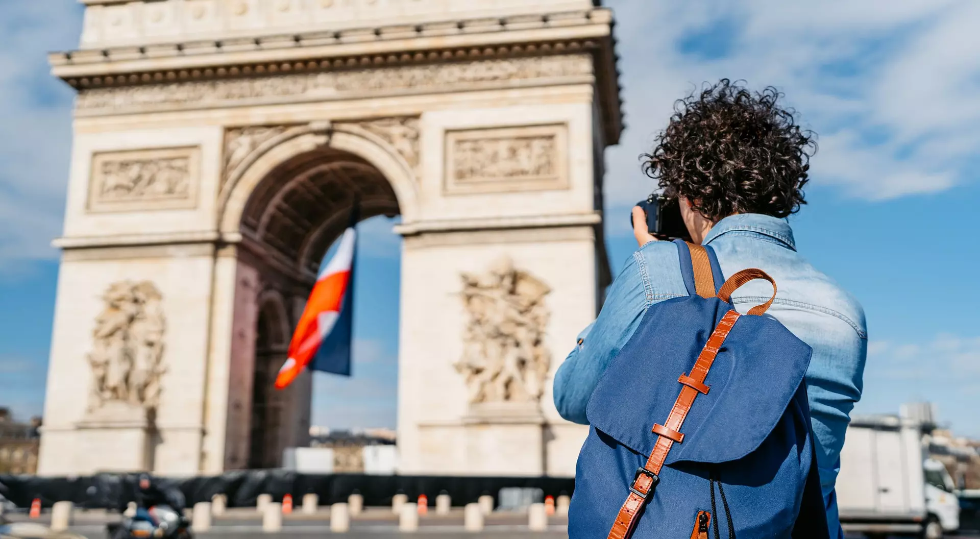 A person with curly hair takes a photo of the Arc de Triomphe, featuring a French flag, under a blue sky.