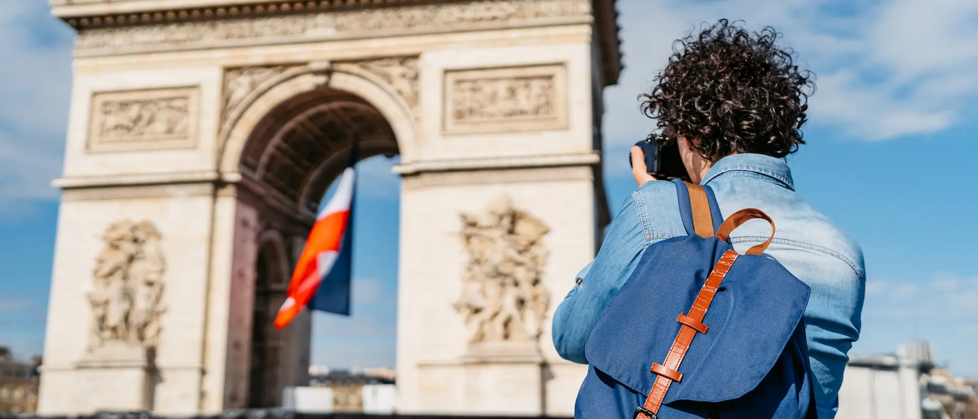 A person with curly hair takes a photo of the Arc de Triomphe, featuring a French flag, under a blue sky.