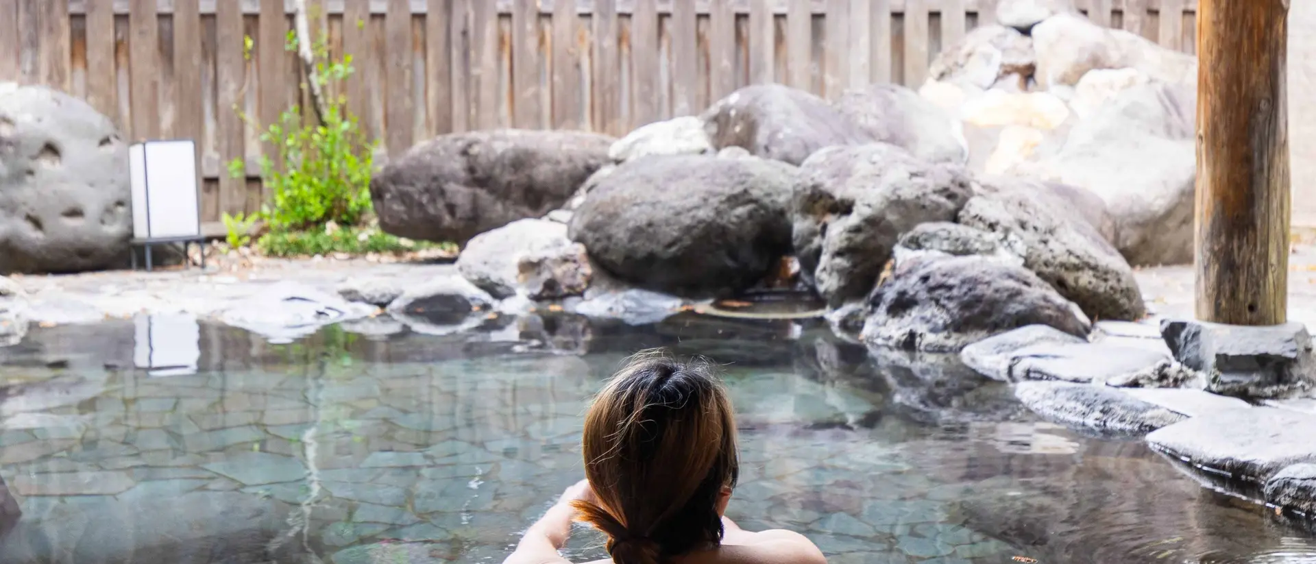 A person with shoulder-length hair relaxes in a stone hot spring surrounded by rocks and greenery.