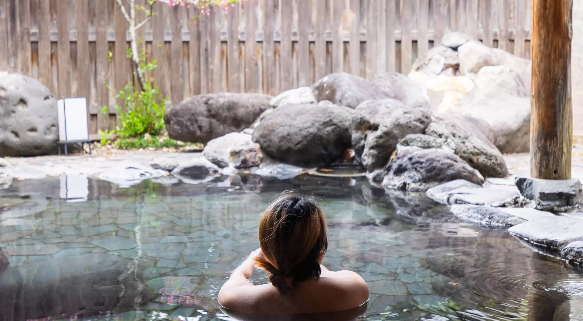A person with shoulder-length hair relaxes in a stone hot spring surrounded by rocks and greenery.