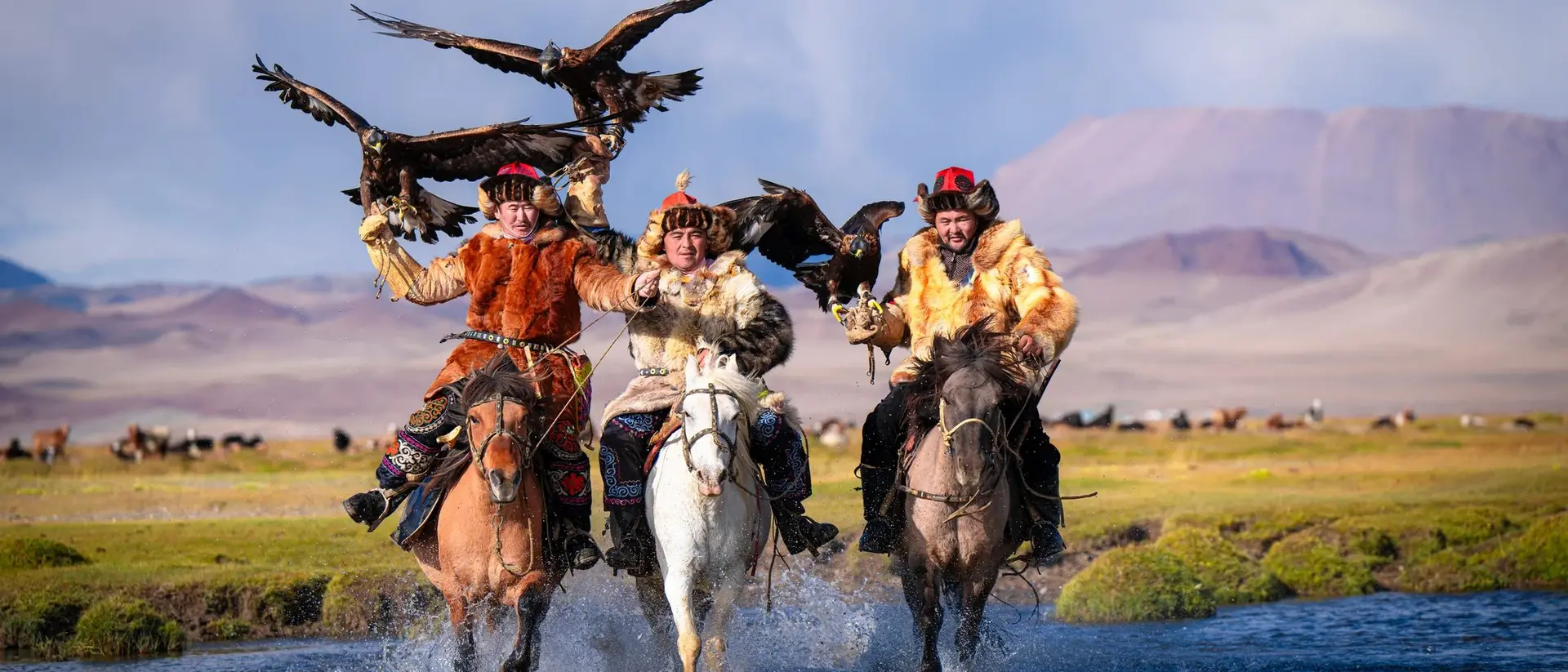 Three riders on horseback splash through a stream, each holding a golden eagle, with mountains in the background.