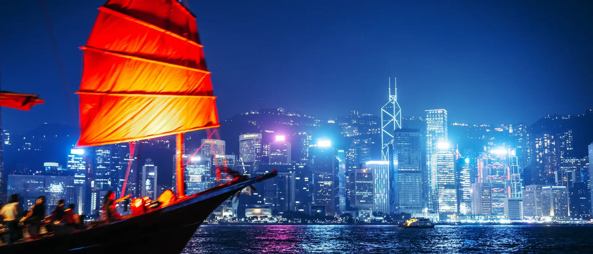 A traditional red-sailed boat in the foreground, with Hong Kong's illuminated skyline in the background at night.