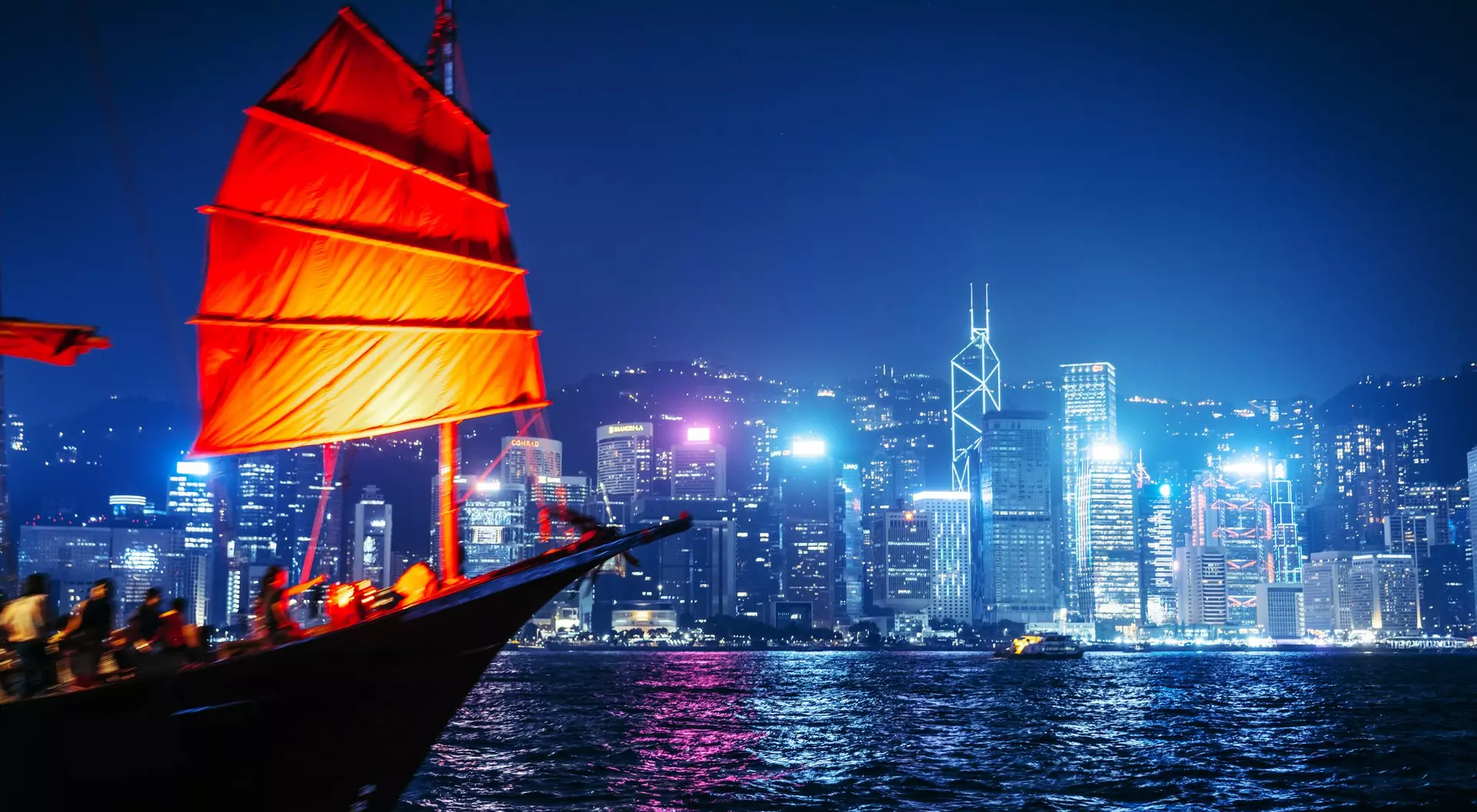 A traditional red-sailed boat in the foreground, with Hong Kong's illuminated skyline in the background at night.