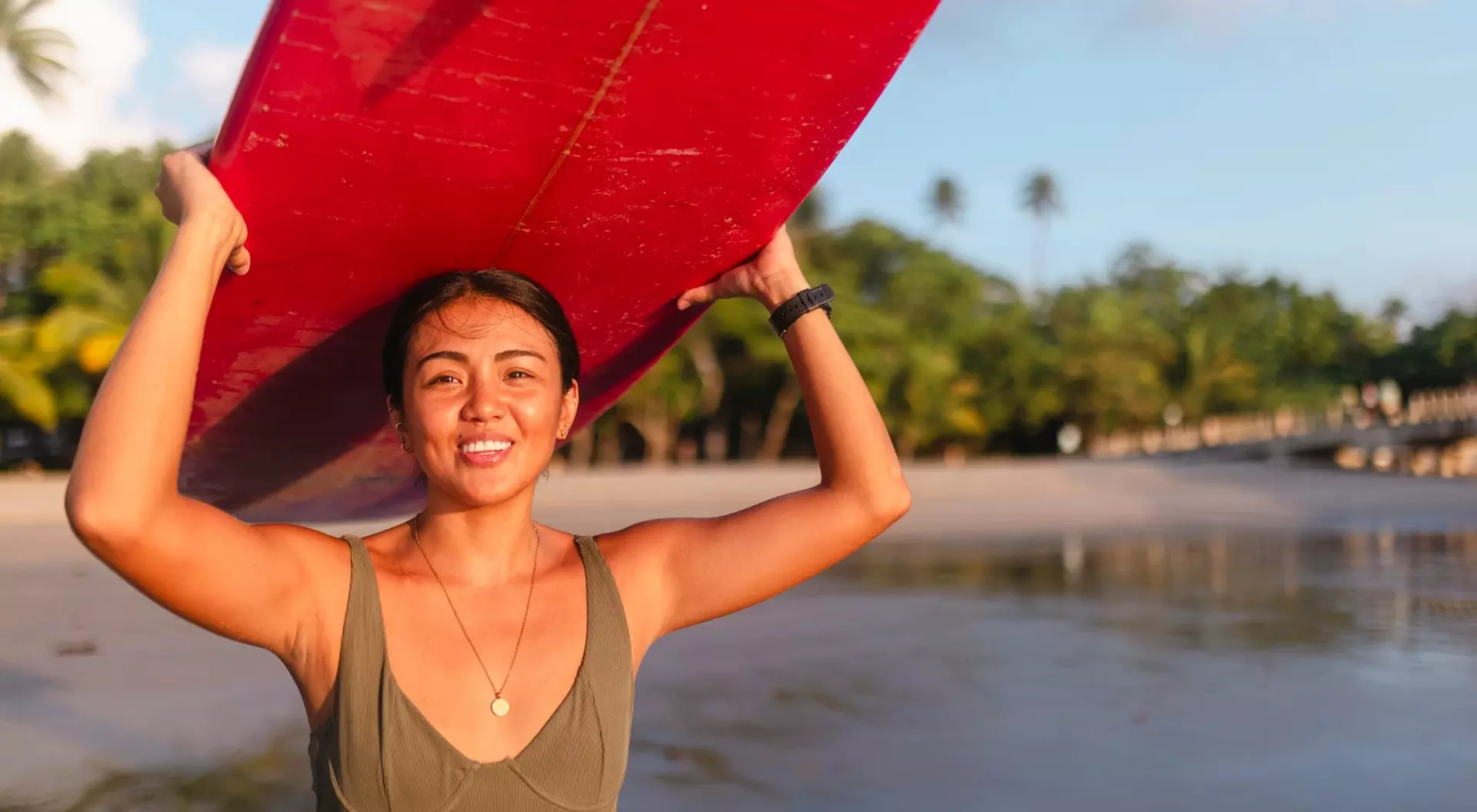 A smiling woman holds a red surfboard overhead at the beach, with palm trees and water in the background.